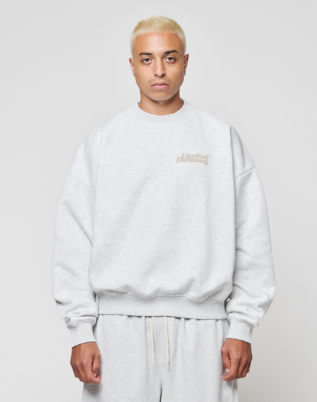 A person with short blond hair wears a LIVE FAST DIE YOUNG Basic Melange Sweater, light gray and oversize fit, standing against a plain background and looking straight at the camera with a neutral expression.