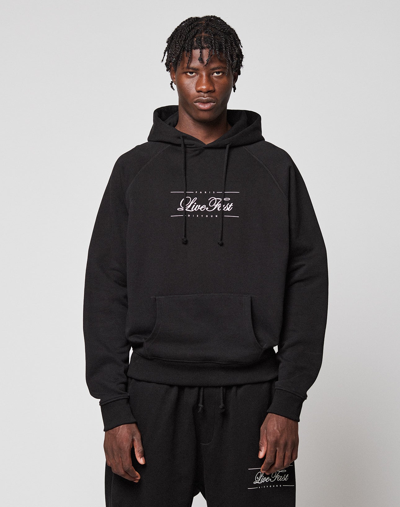 A man with short, textured hair wears a black LFDY Embroidery Hooded hoodie and matching sweatpants by LIVE FAST DIE YOUNG, both with white Clive Rest logos. He stands against a plain light gray background, looking directly at the camera. | model