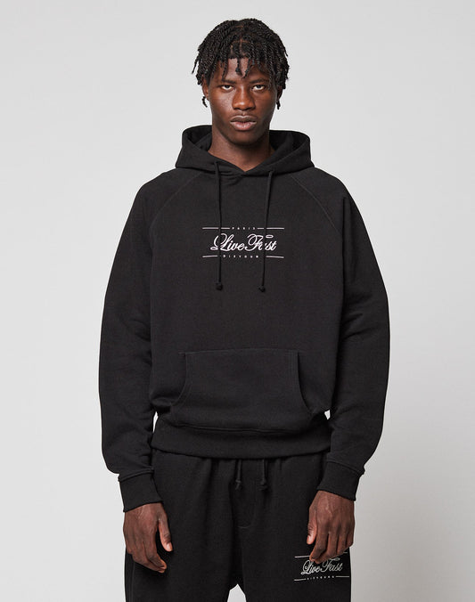 A man with short, textured hair wears a black LFDY Embroidery Hooded hoodie and matching sweatpants by LIVE FAST DIE YOUNG, both with white Clive Rest logos. He stands against a plain light gray background, looking directly at the camera. | model