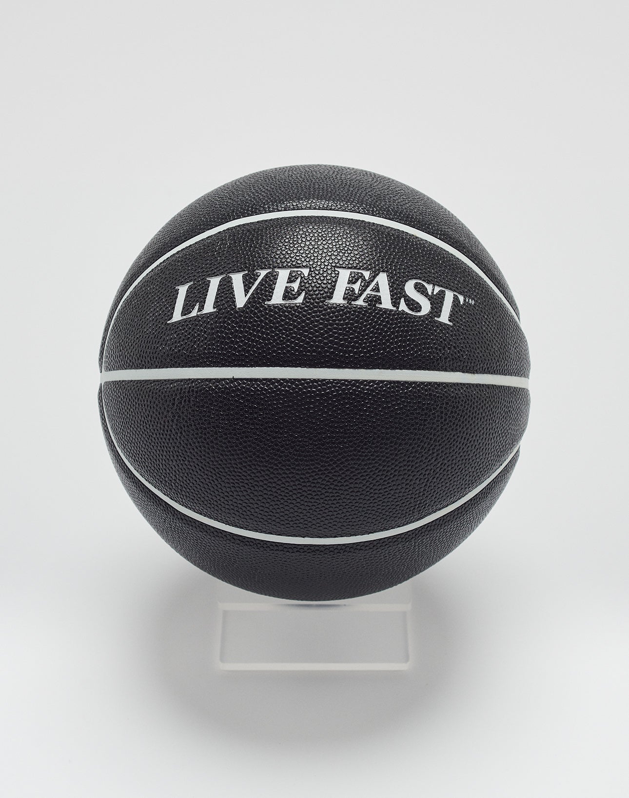 The Live Fast Basketball by LIVE FAST DIE YOUNG features a black design with white stripes, deep grooves, and LIVE FAST branding in hygroscopic PU, displayed on a clear stand against a white background.