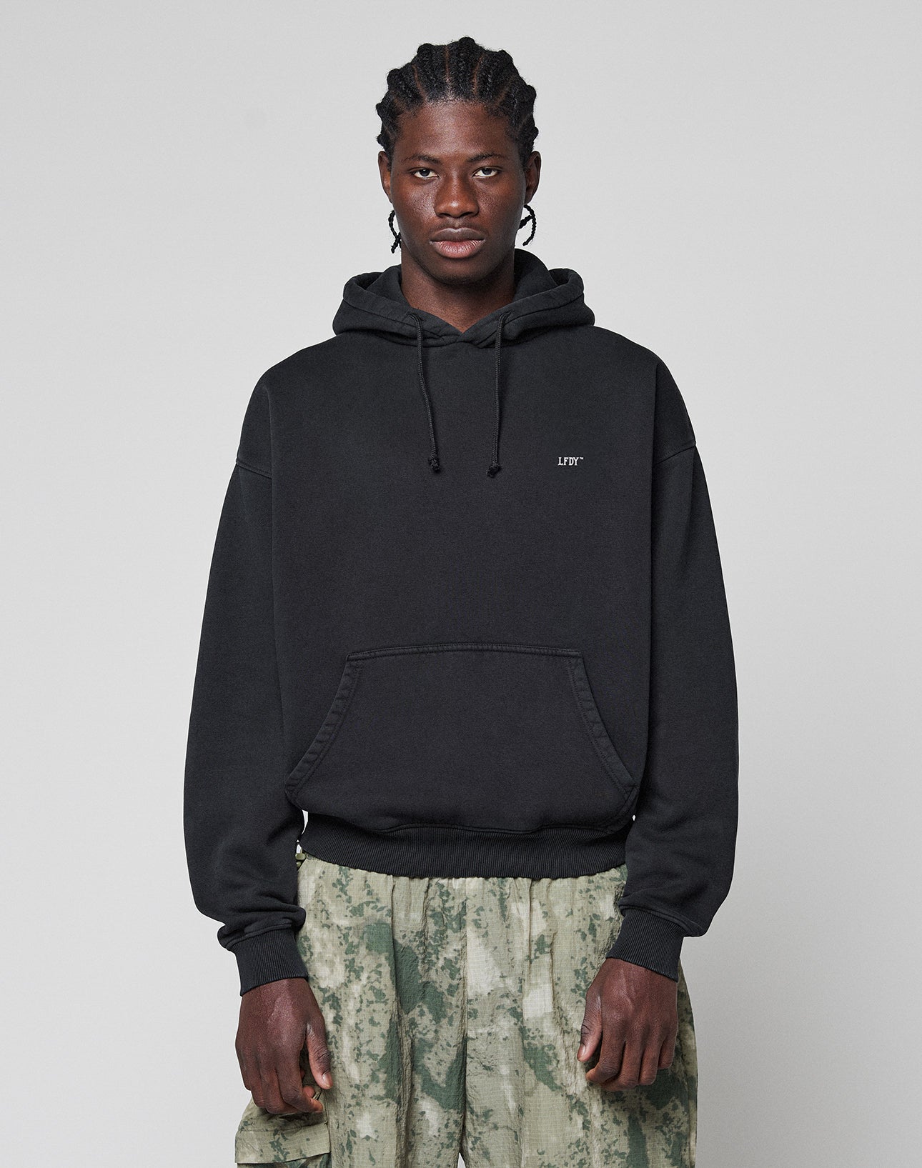 A person with braided hair wears the Live Fast Vegas Hooded by LIVE FAST DIE YOUNG, featuring LIFE on the chest and front/back prints. The black hoodie (65% cotton) is styled with light green patterned pants against a gray background.