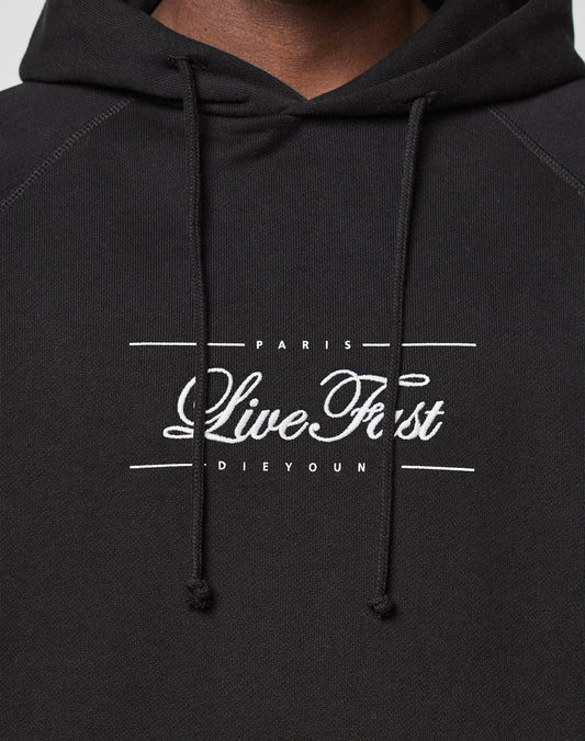 A close-up of the black LFDY Embroidery Hooded by LIVE FAST DIE YOUNG, made from 100% cotton French terry, with visible hood drawstrings and “PARIS Live Fast DIE YOUNG” white text embroidery on the chest.