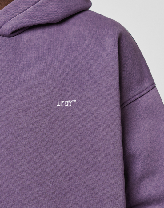 Close-up of a person wearing the Basic Evidence Hooded by LIVE FAST DIE YOUNG, a purple oversize hoodie with small white LFDY™ logo on the chest. Made from Baumwolle Polyester, shown partially against a plain background.