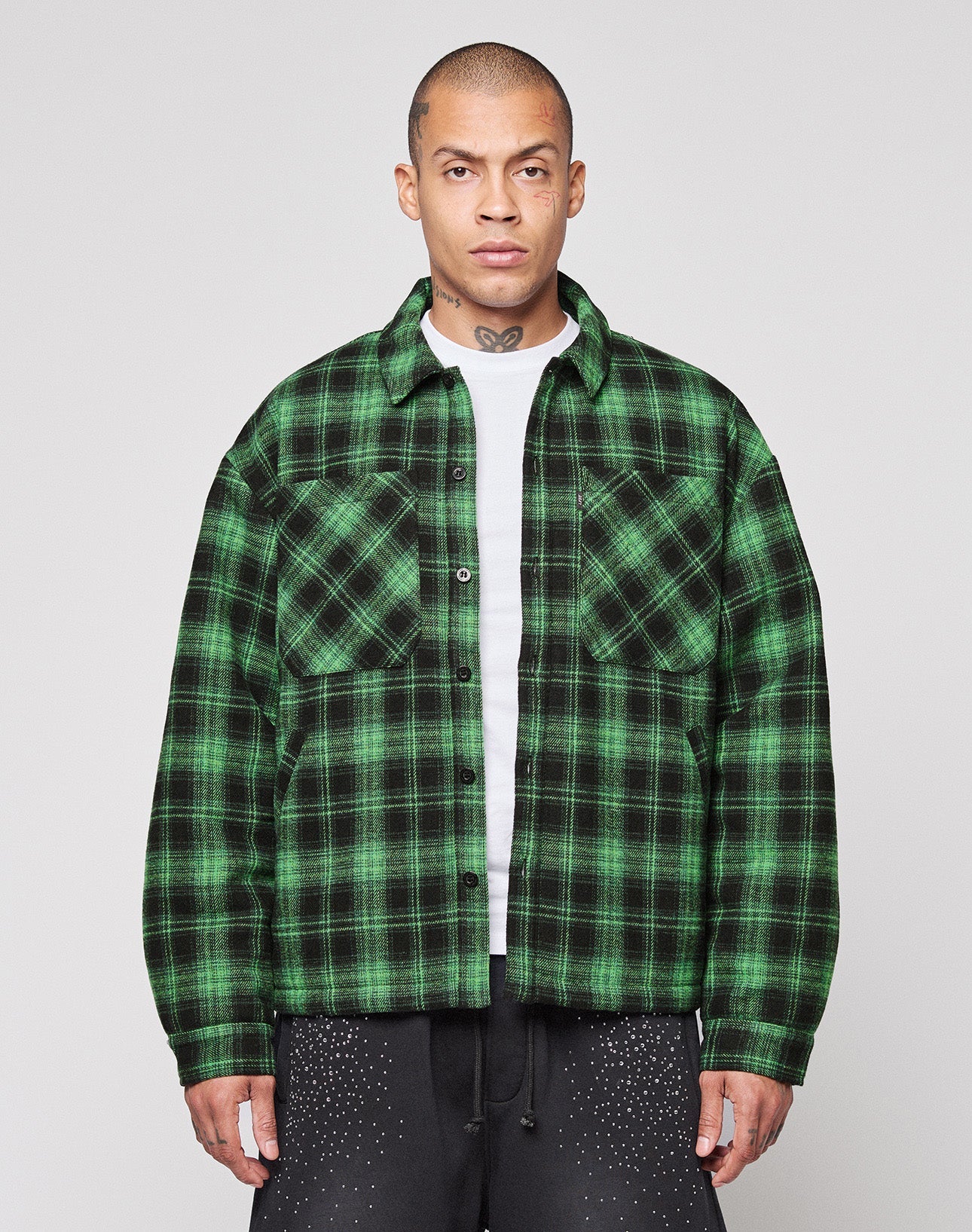 A person stands facing forward in a LIVE FAST DIE YOUNG LFDY Padded Flannel—green and black plaid, worn over a white T-shirt with black pants with white speckles. The background is plain and light-colored. | model