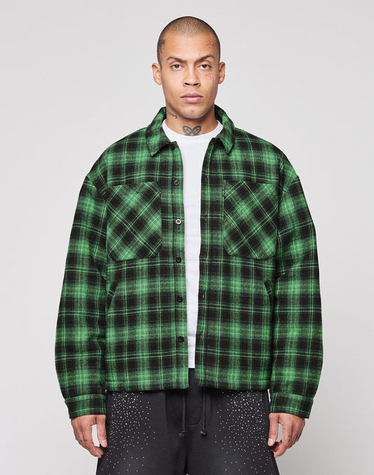 A person stands facing forward in a LIVE FAST DIE YOUNG LFDY Padded Flannel—green and black plaid, worn over a white T-shirt with black pants with white speckles. The background is plain and light-colored. | model