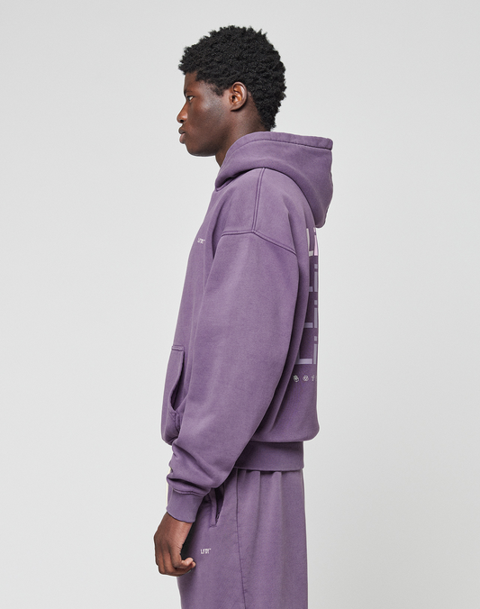 A person stands in profile, hands at their sides, wearing the LIVE FAST DIE YOUNG Basic Evidence Hooded purple oversize-fit hoodie and matching sweatpants against a plain light background.