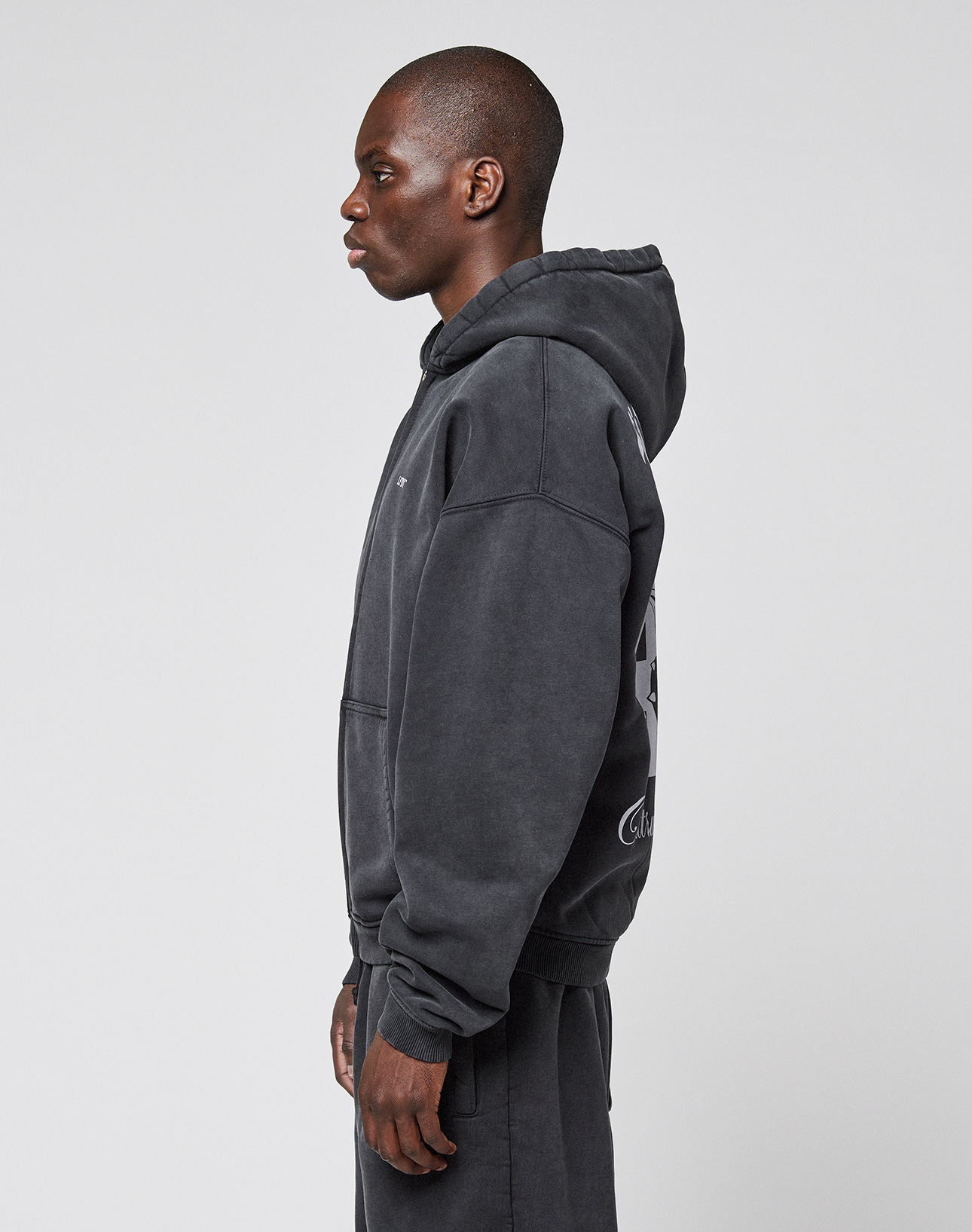 A person in profile wears the LFDY Run Club Hooded Zip by LIVE FAST DIE YOUNG, styled with matching pants. The dark gray oversize fit, soft Baumwolle-Polyester blend, and subtle Ärmelprint lend a modern touch to their casual look.