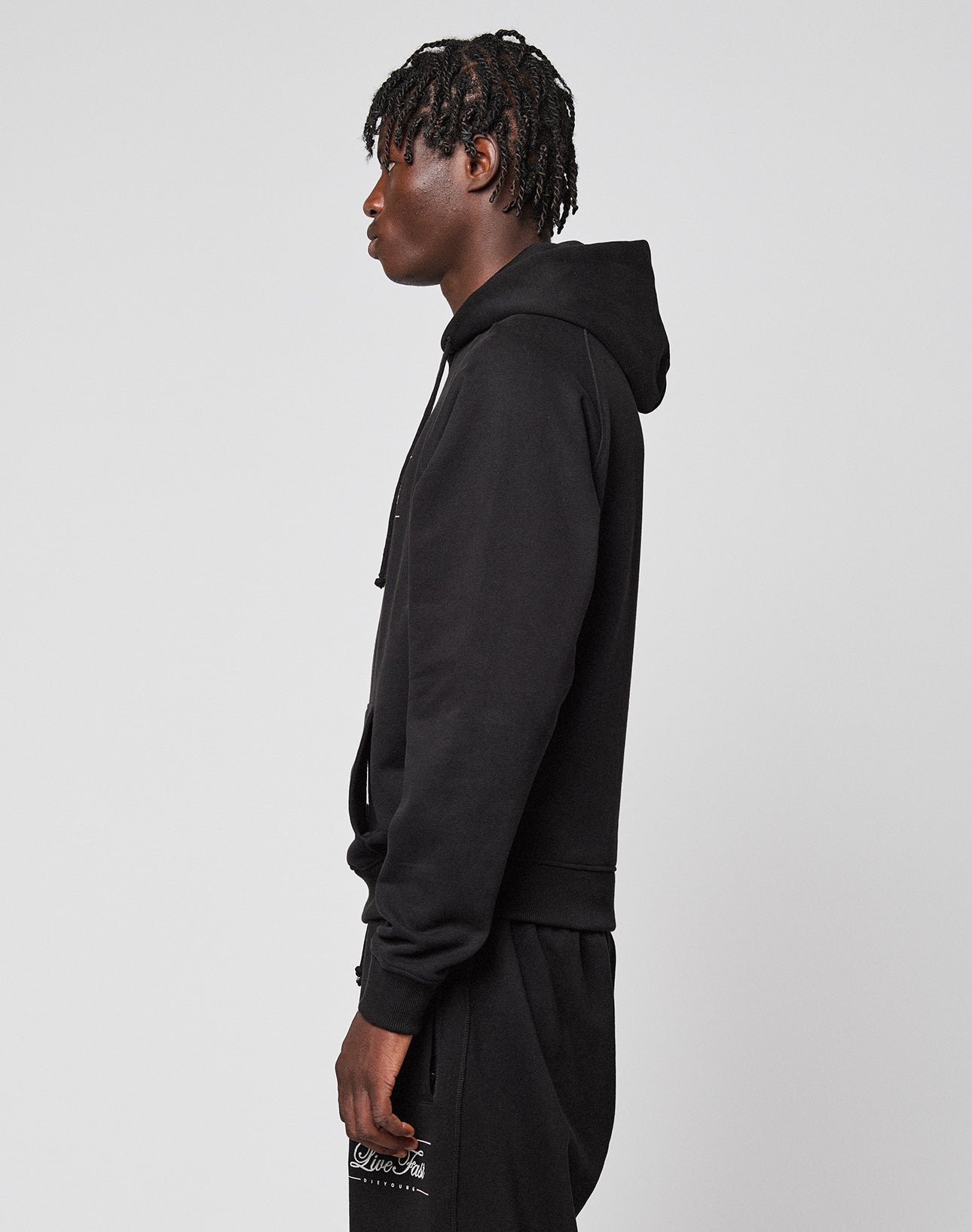 A person with short dreadlocks stands in profile, facing left, wearing a black LFDY Embroidery Hooded by LIVE FAST DIE YOUNG and matching black pants against a plain light background.
