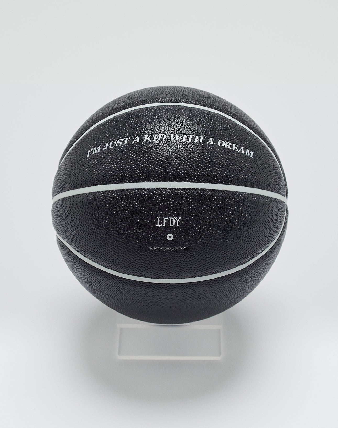 A Live Fast Basketball from LIVE FAST DIE YOUNG, black with white stripes and deep grooves, sits on a clear stand. It features IM JUST A KID WITH A DREAM, the initials LFDY, and small extra text below.