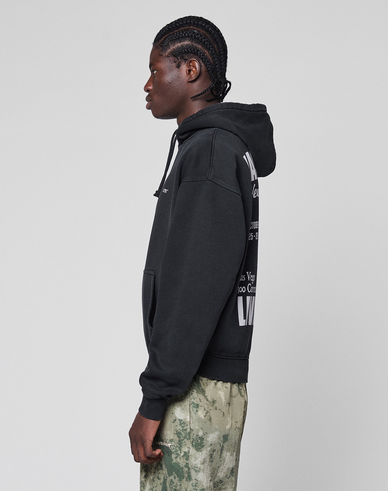 A person with braided hair stands in profile, wearing the LIVE FAST DIE YOUNG Live Fast Vegas Hooded in an oversize fit with front and back prints, paired with white and green camouflage pants (65% Baumwolle), against a light gray background.