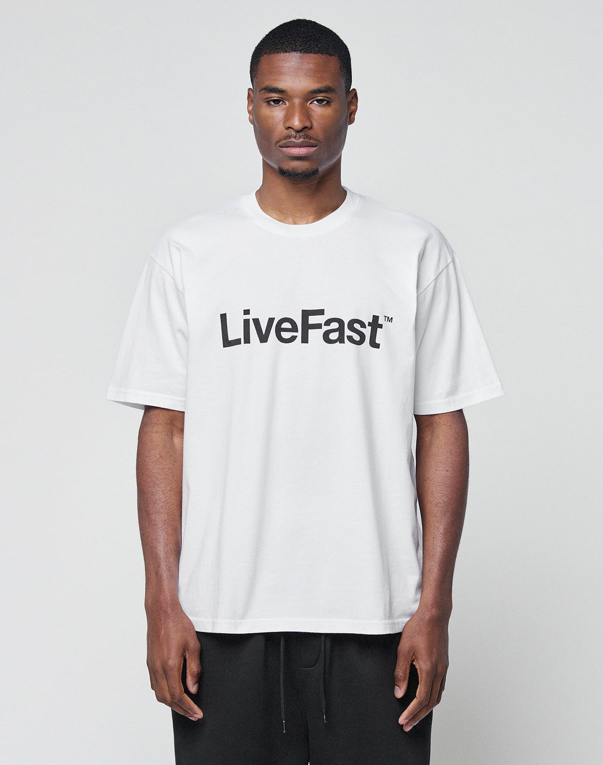 A man faces forward, wearing the Basic LiveFast Tee by LIVE FAST DIE YOUNG, a loose white T-shirt with LiveFast in bold black letters on the chest. The Regular Fit shirt is photographed against a plain, light background. | model