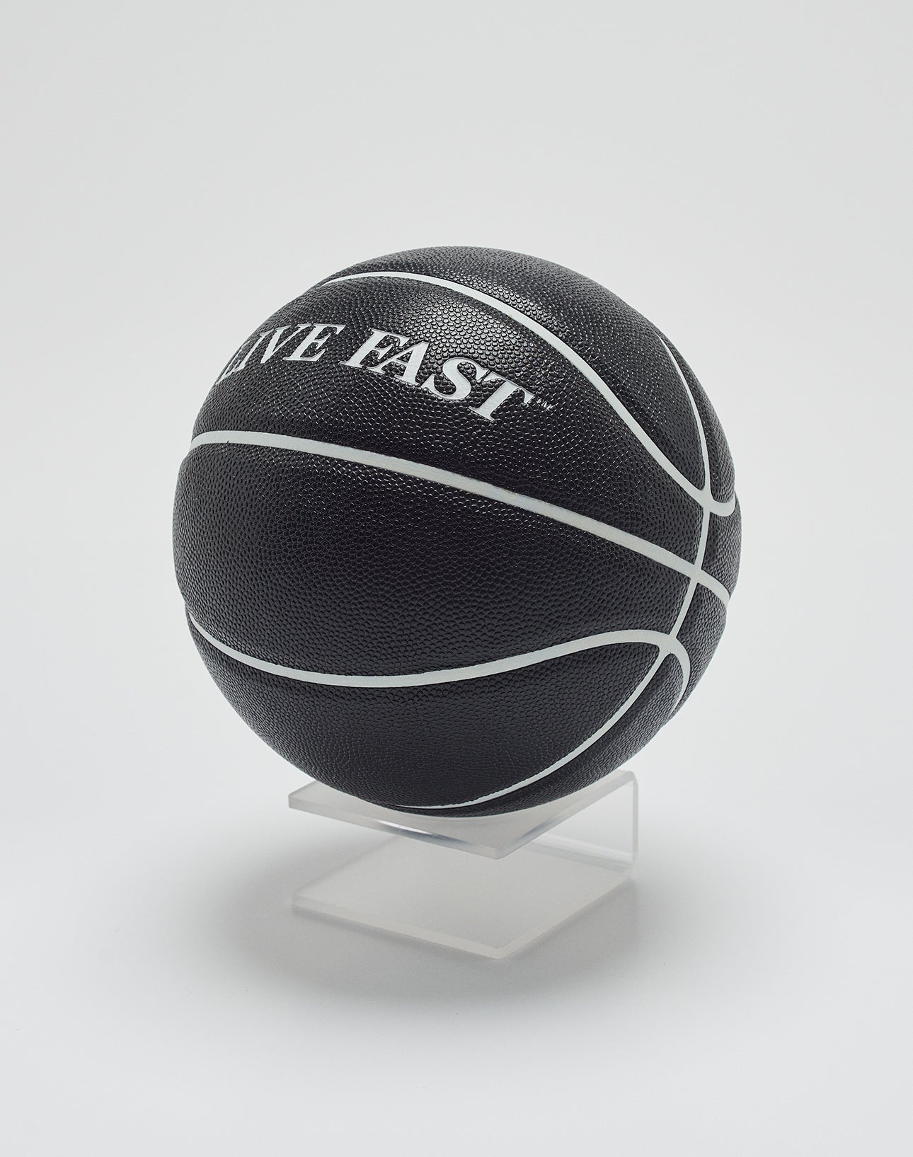 The Live Fast Basketball by LIVE FAST DIE YOUNG is black with deep grooves and white lines, featuring LIVE FAST branding. It is displayed on a clear stand against a plain white background.
