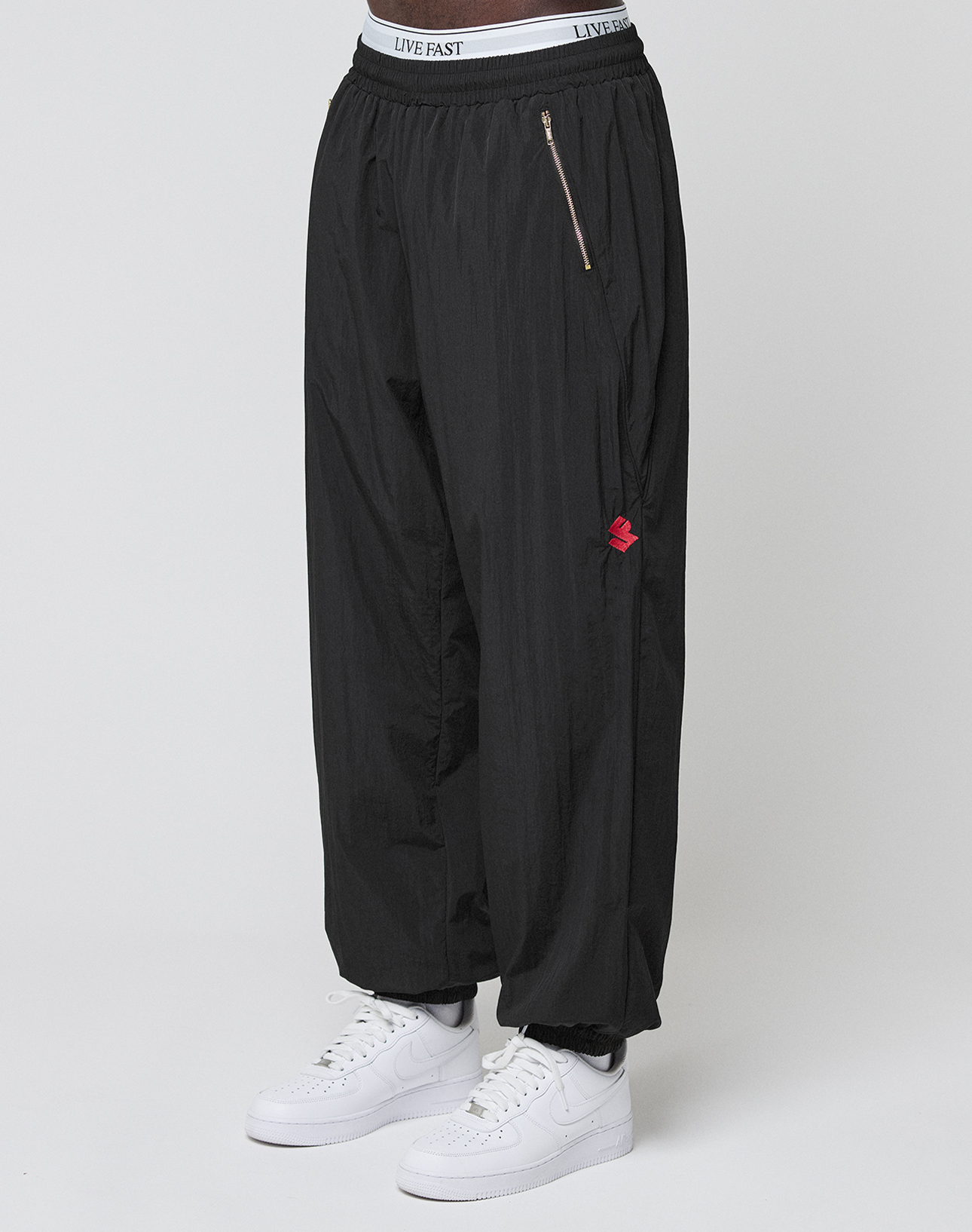 A person stands against a plain background wearing LFDY Dealership Trackpants by LIVE FAST DIE YOUNG—loose black track pants with an elastic waistband labeled LIVE FAST and a zipper pocket—paired with white Nike sneakers. | model