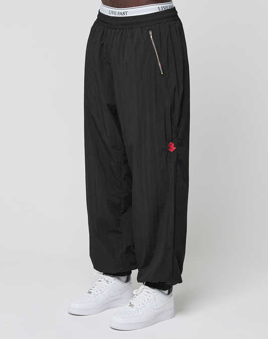 A person stands against a plain background wearing LFDY Dealership Trackpants by LIVE FAST DIE YOUNG—loose black track pants with an elastic waistband labeled LIVE FAST and a zipper pocket—paired with white Nike sneakers. | model