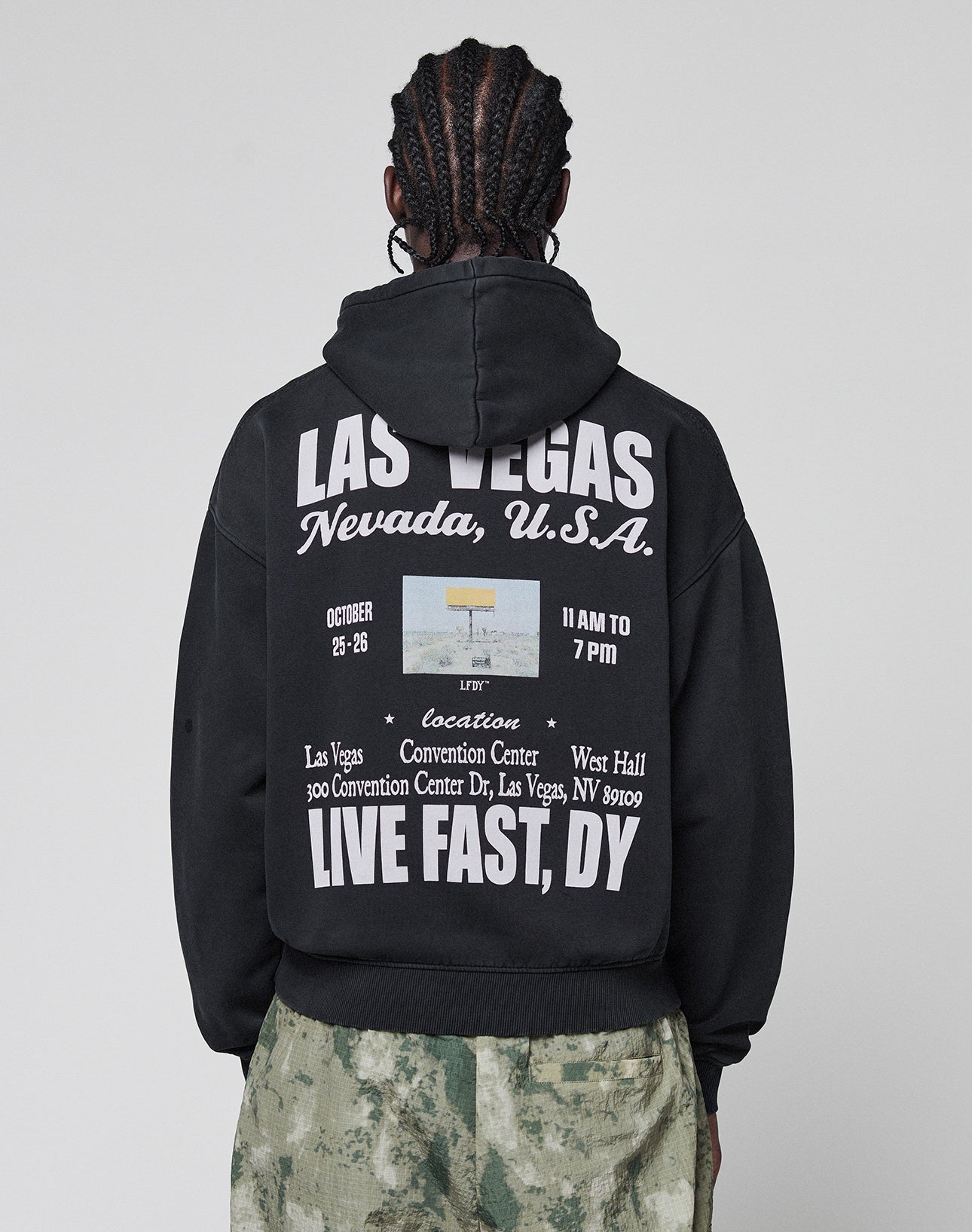 A person with braided hair wears the LIVE FAST DIE YOUNG Live Fast Vegas Hooded with white front and back print plus a small photo for an event at the Las Vegas Convention Center, paired with green camouflage pants. | model