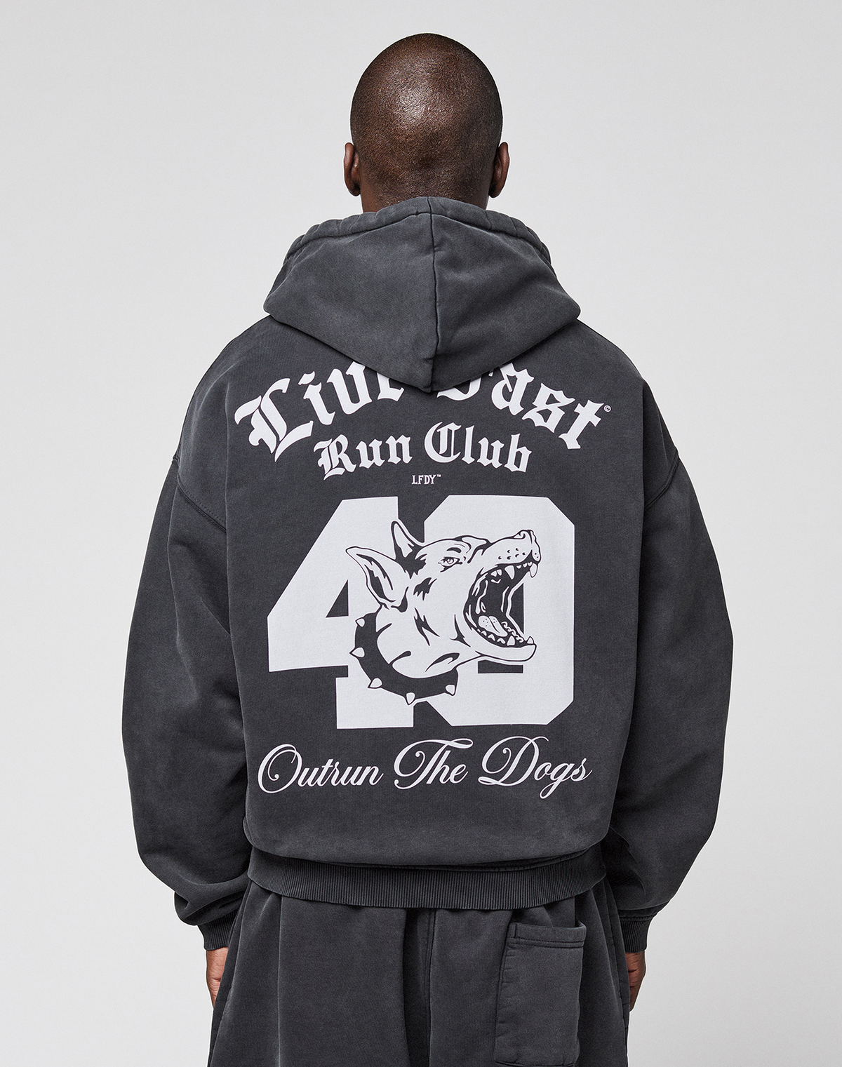 Someone wearing the LIVE FAST DIE YOUNG LFDY Run Club Hooded Zip, a black oversized hoodie featuring bold white graphics on the back: "Live Fast Run Club," "49," "Outrun The Dogs," and a snarling dog head. Made from cotton-polyester. | model