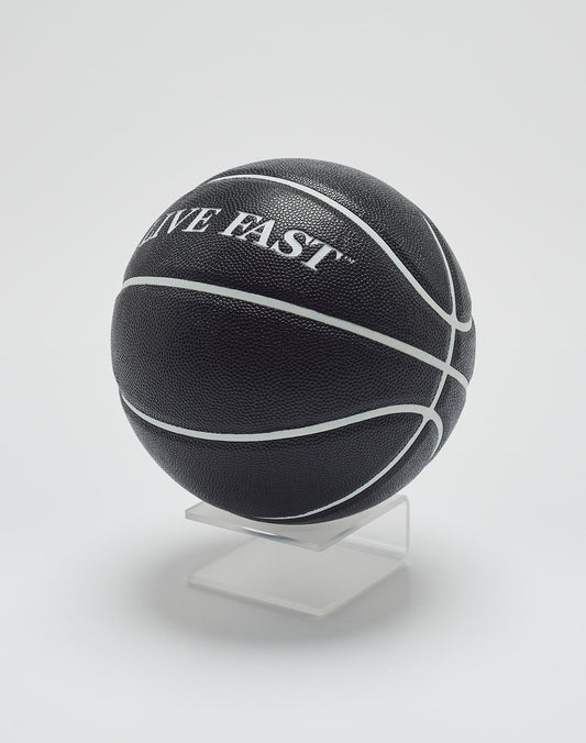 The Live Fast Basketball by LIVE FAST DIE YOUNG is black with deep grooves and white lines, featuring LIVE FAST branding. It is displayed on a clear stand against a plain white background.