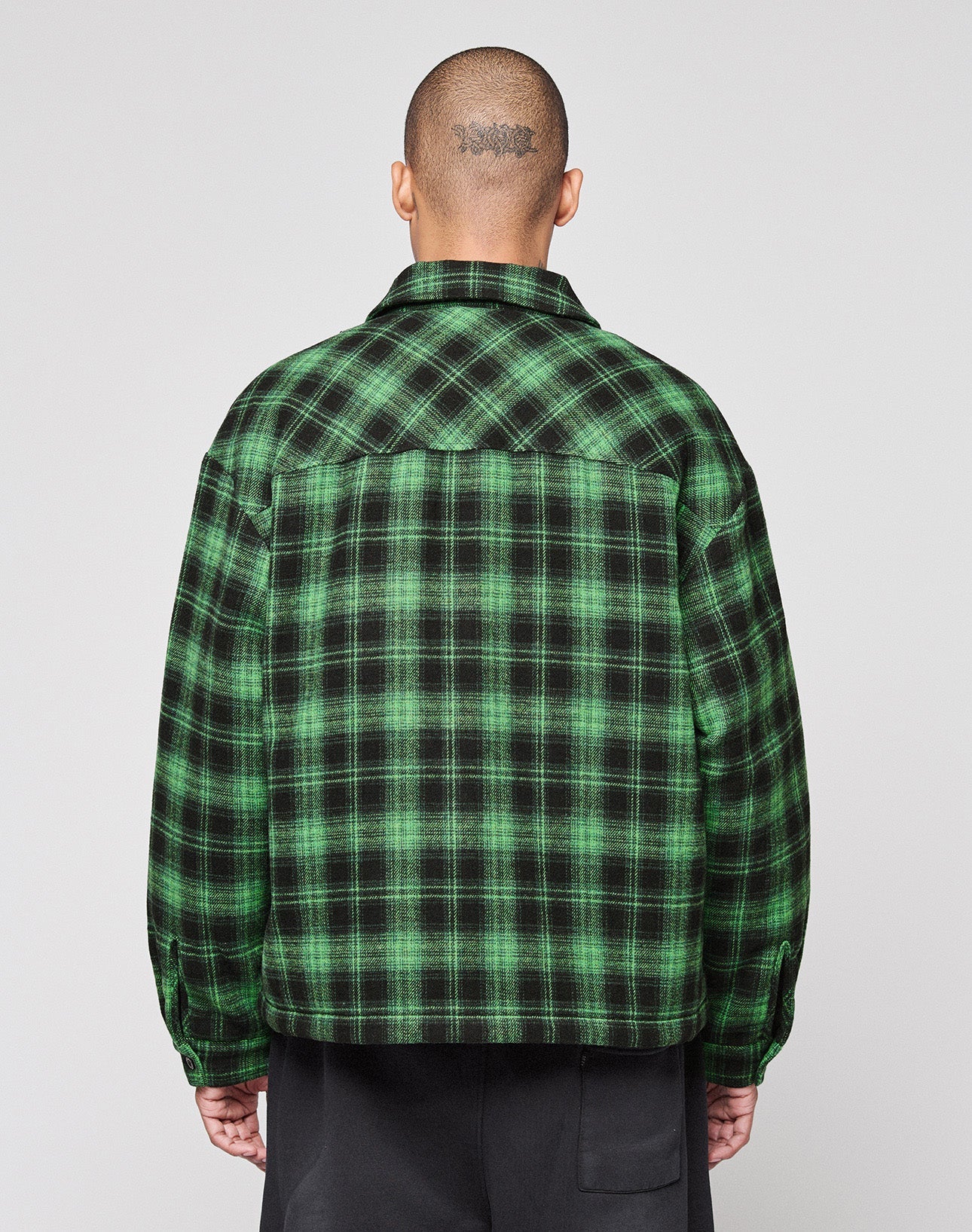 A person with a shaved head and visible tattoo faces away, wearing a green and black LIVE FAST DIE YOUNG LFDY Padded Flannel and black pants against a plain light background.