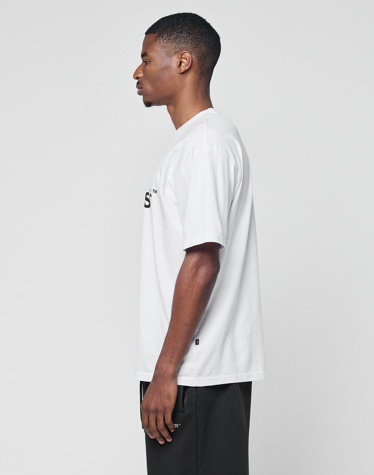 A man stands in profile against a plain background, wearing the LIVE FAST DIE YOUNG Basic LiveFast Tee in Regular Fit and dark pants.