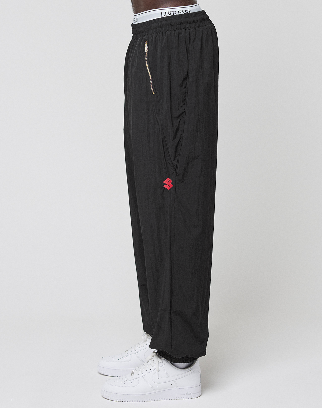 A person stands sideways in LIVE FAST DIE YOUNG’s LFDY Dealership Trackpants—black, wide fit, with a red logo and visible LIVE FAST elastic waistband—paired with white sneakers against a plain white background.