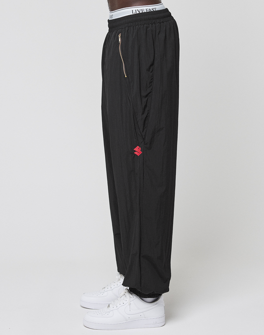 A person stands sideways in LIVE FAST DIE YOUNG’s LFDY Dealership Trackpants—black, wide fit, with a red logo and visible LIVE FAST elastic waistband—paired with white sneakers against a plain white background.