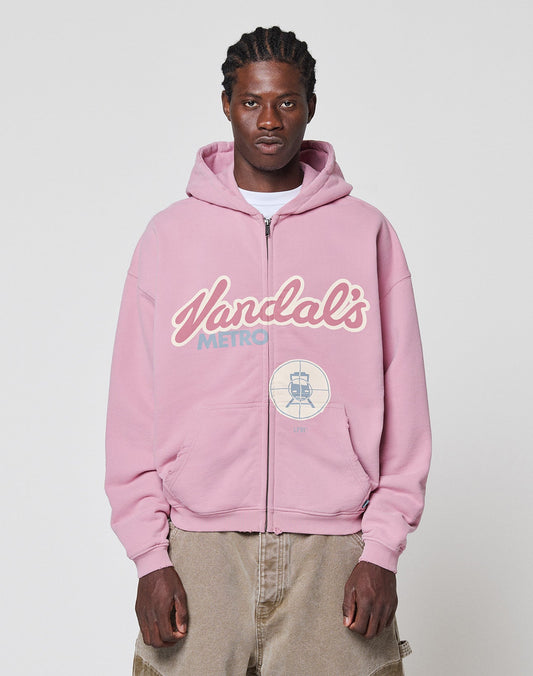 A man faces the camera in a neutral pose, wearing the LIVE FAST DIE YOUNG LFDY Vandals Hooded Zip in light pink with a “Vandals Metro” front print, paired with beige pants, against a plain background. | model