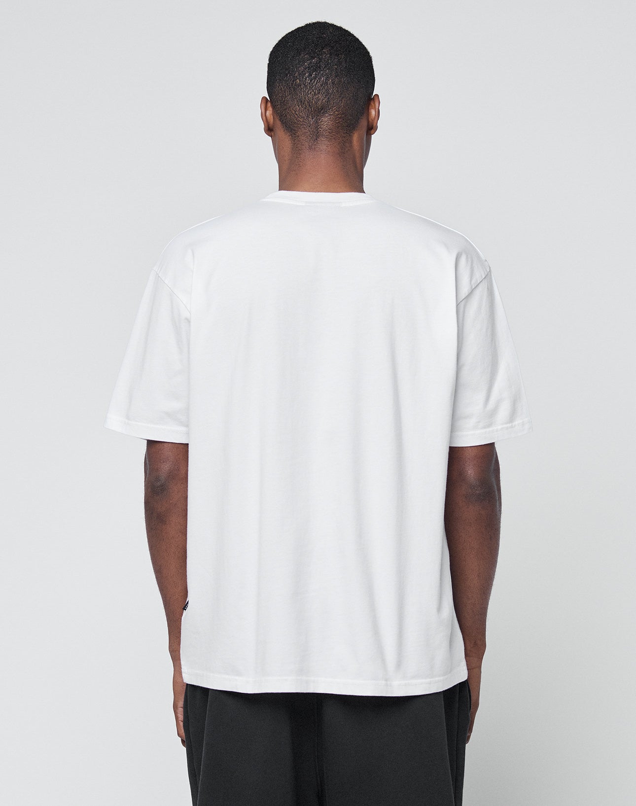A person faces away from the camera wearing the LIVE FAST DIE YOUNG Basic LiveFast Tee, a plain white regular-fit short-sleeve shirt, with black pants against a neutral gray background.
