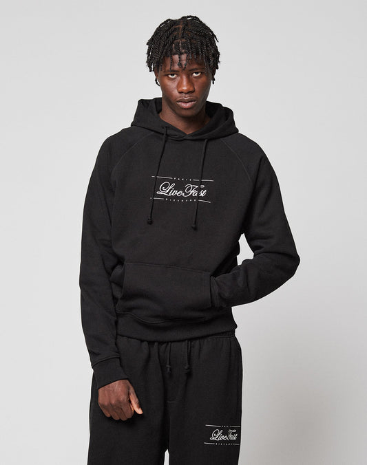 A young man with short braids models the LIVE FAST DIE YOUNG LFDY Embroidery Hooded and matching black sweatpants with white LiveVäst text, made from soft French terry, standing against a plain light gray background.
