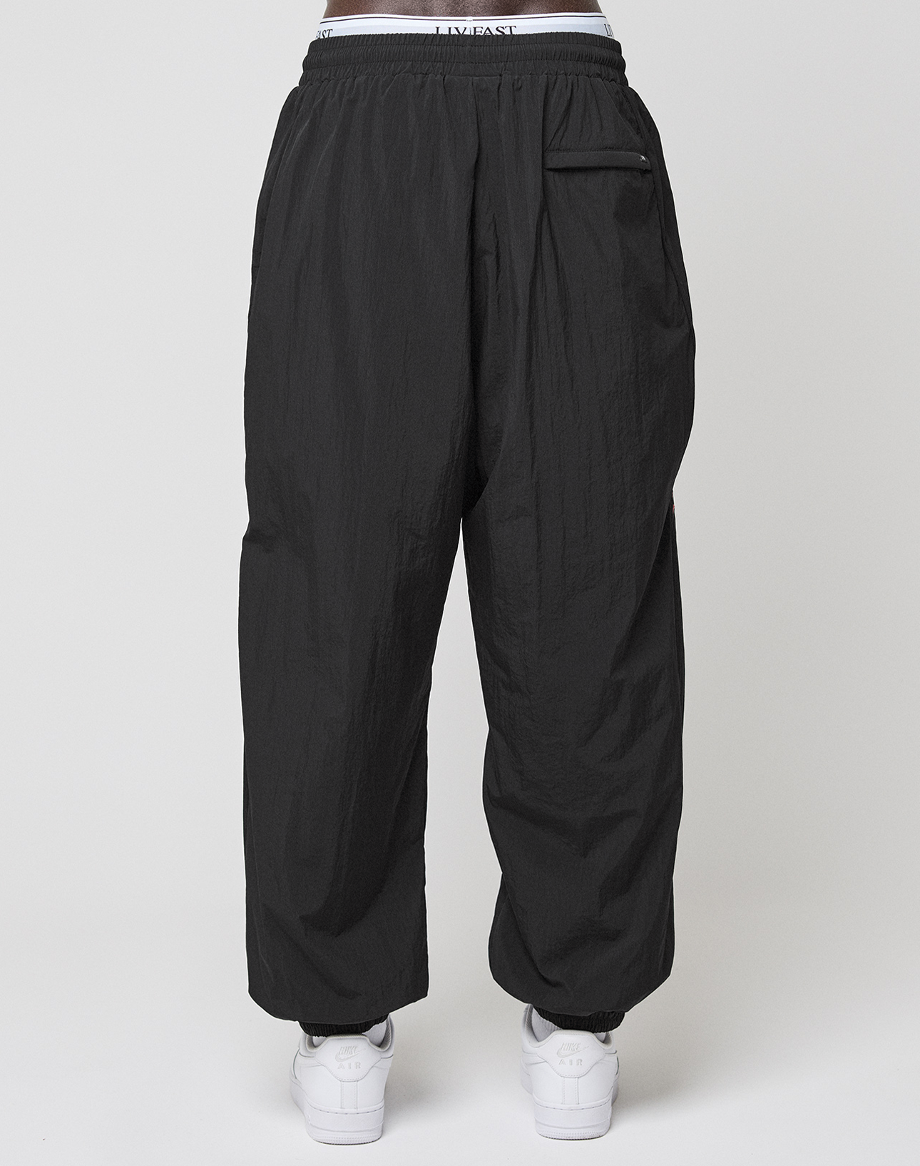 A person wears LIVE FAST DIE YOUNG LFDY Dealership Trackpants in black polyamide with a relaxed wide fit, elastic waistband, and a single back pocket, paired with white sneakers. Shown from the back against a plain light background.