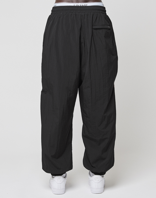 A person wears LIVE FAST DIE YOUNG LFDY Dealership Trackpants in black polyamide with a relaxed wide fit, elastic waistband, and a single back pocket, paired with white sneakers. Shown from the back against a plain light background.