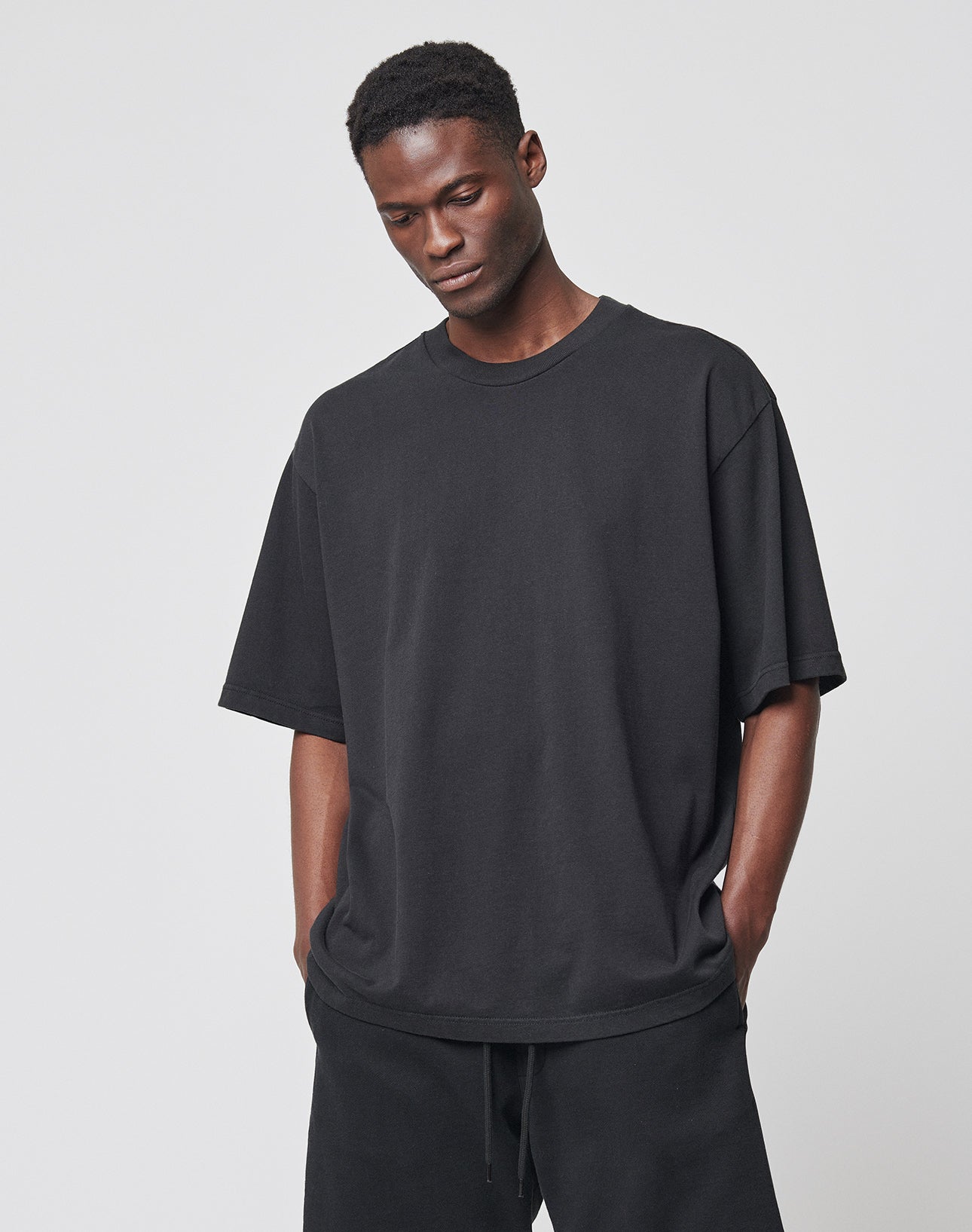 A man stands against a light gray background, hands in his pockets, wearing the LIVE FAST DIE YOUNG Basic Heavy Tee in black with matching black pants, looking down.