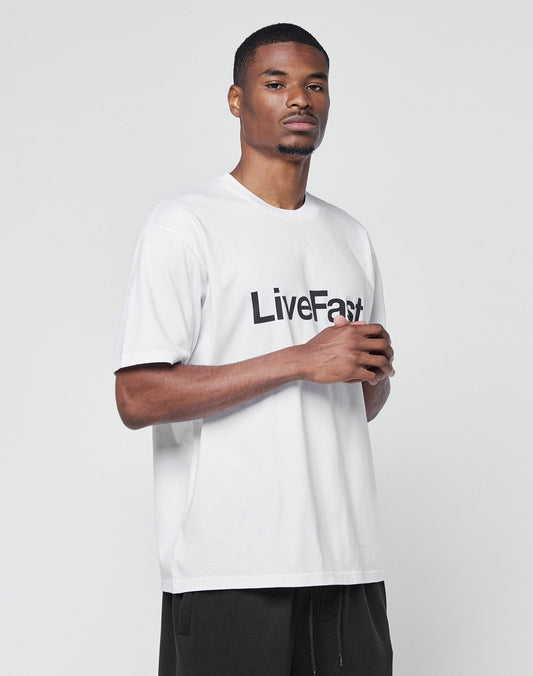 A man stands against a white background wearing the LIVE FAST DIE YOUNG Basic LiveFast Tee—a regular fit white t-shirt with “LiveFast” in bold black letters—and black pants, looking neutrally at the camera.