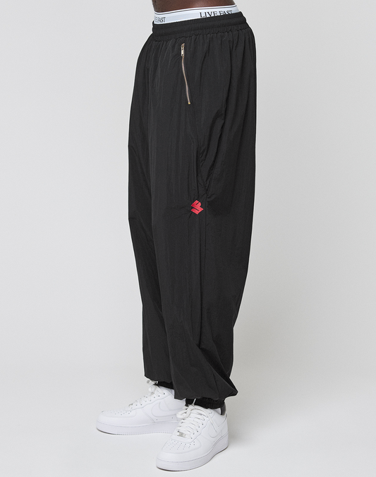 A person wears LFDY Dealership Trackpants by LIVE FAST DIE YOUNG—wide fit, black, with a zipper pocket, small red logo, visible white “LIVE FAST” waistband—and white sneakers, standing against a plain white background.