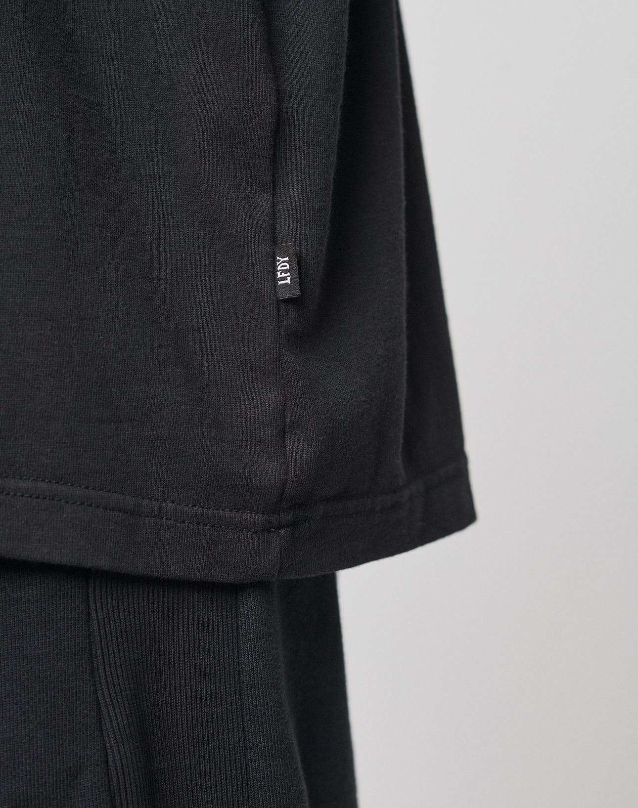 Close-up of the lower side of a Basic Heavy Tee by LIVE FAST DIE YOUNG in black Baumwolle, showing a small black ART tag near the seam. The oversize tee is worn over ribbed black pants against a light plain background.