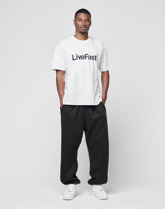 A man faces forward in a white LIVE FAST DIE YOUNG Basic LiveFast Tee with LiveFast in black, paired with loose black pants and white sneakers, against a plain light background.