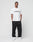 A man faces forward in a white LIVE FAST DIE YOUNG Basic LiveFast Tee with LiveFast in black, paired with loose black pants and white sneakers, against a plain light background.
