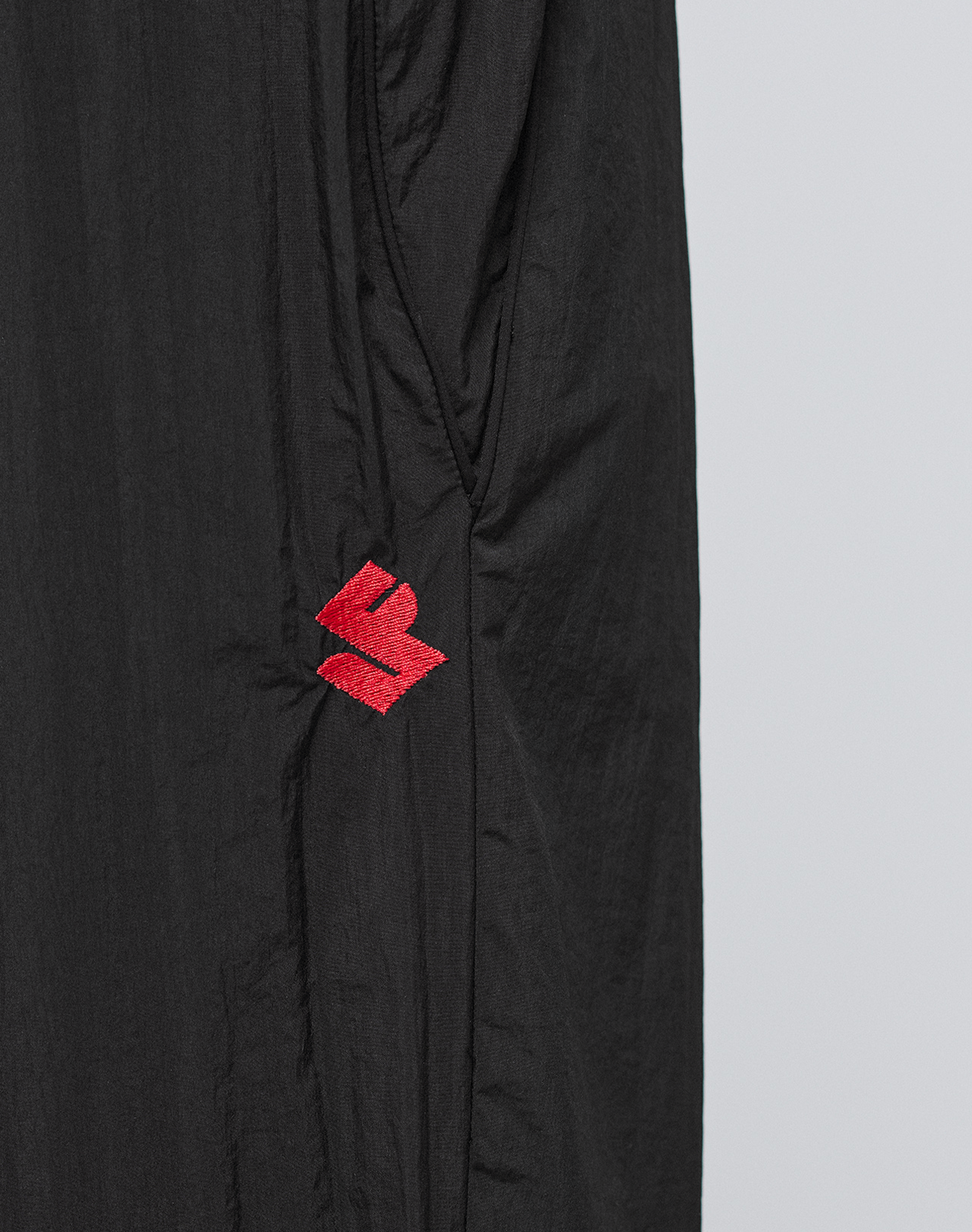 Close-up of lightweight, slightly crinkled polyamid fabric on the LFDY Dealership Trackpants by LIVE FAST DIE YOUNG, featuring a small red abstract embroidered logo near the left side seam.