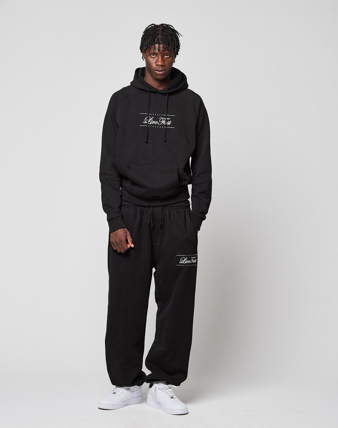A person stands against a plain background in a loose black hoodie and LIVE FAST DIE YOUNG Wide Cuff Joggers, both with white text. White sneakers and short dreadlocks complete their relaxed look.