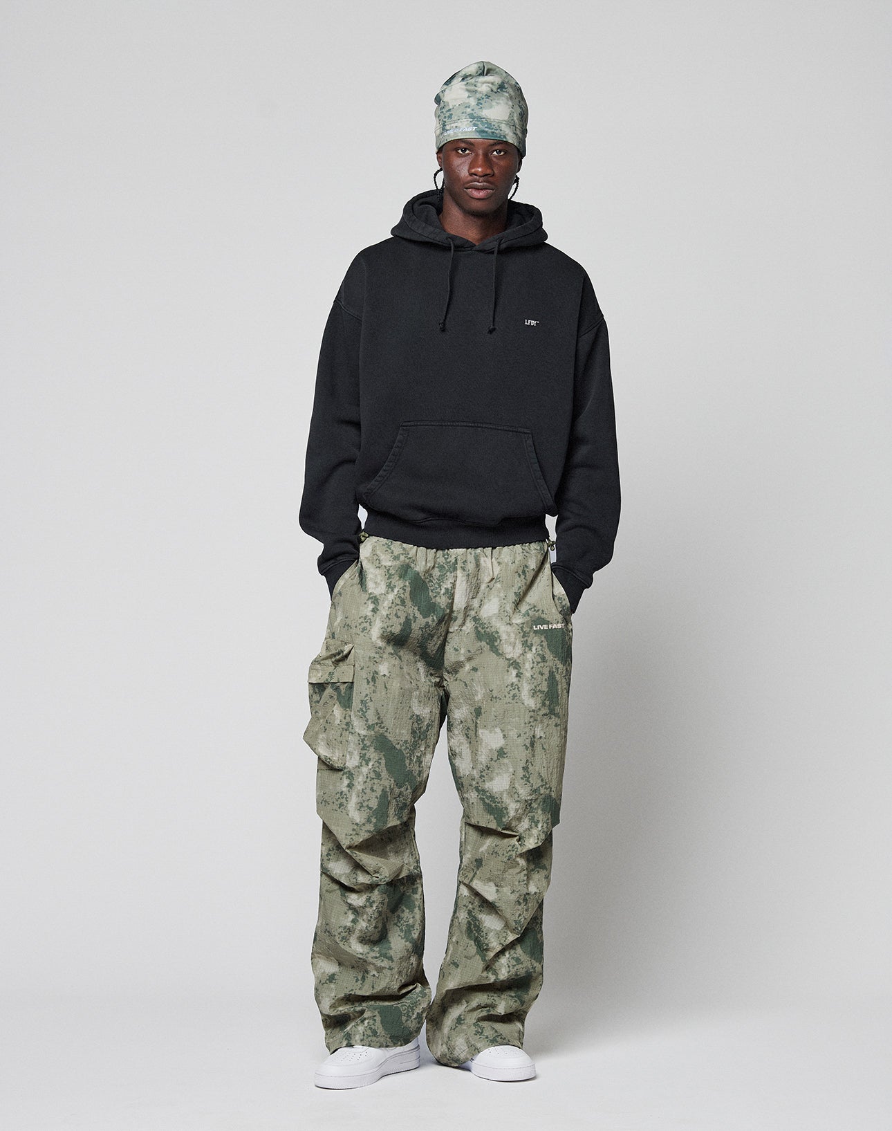 A person stands facing forward, wearing the Live Fast Vegas Hooded by LIVE FAST DIE YOUNG (65% Baumwolle), green camo cargo pants, a matching green headscarf, and white sneakers against a plain light gray background.