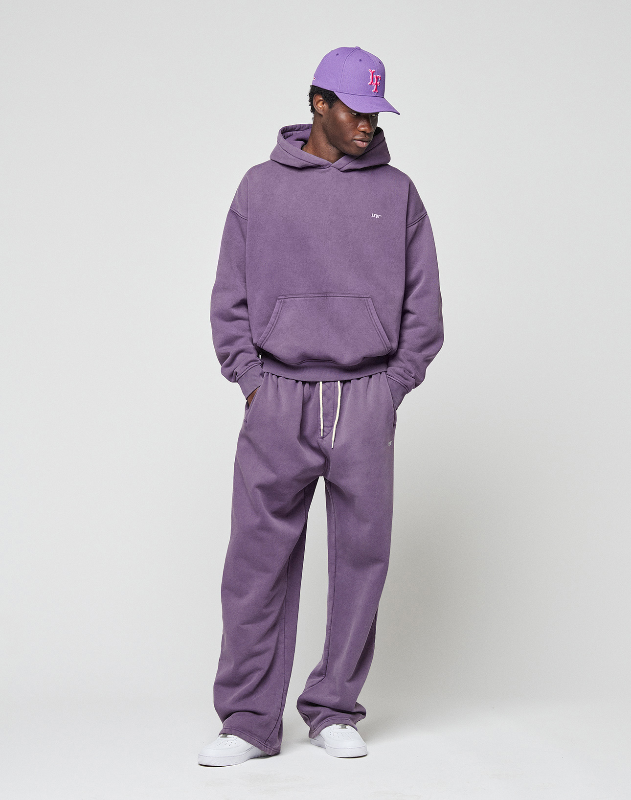 A person wearing a purple LIVE FAST DIE YOUNG Basic Wide Jogger set with an elastic waistband, a matching purple baseball cap, and white sneakers stands against a plain light background.