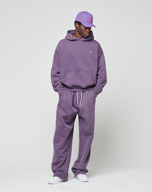 A person wearing a purple LIVE FAST DIE YOUNG Basic Wide Jogger set with an elastic waistband, a matching purple baseball cap, and white sneakers stands against a plain light background.