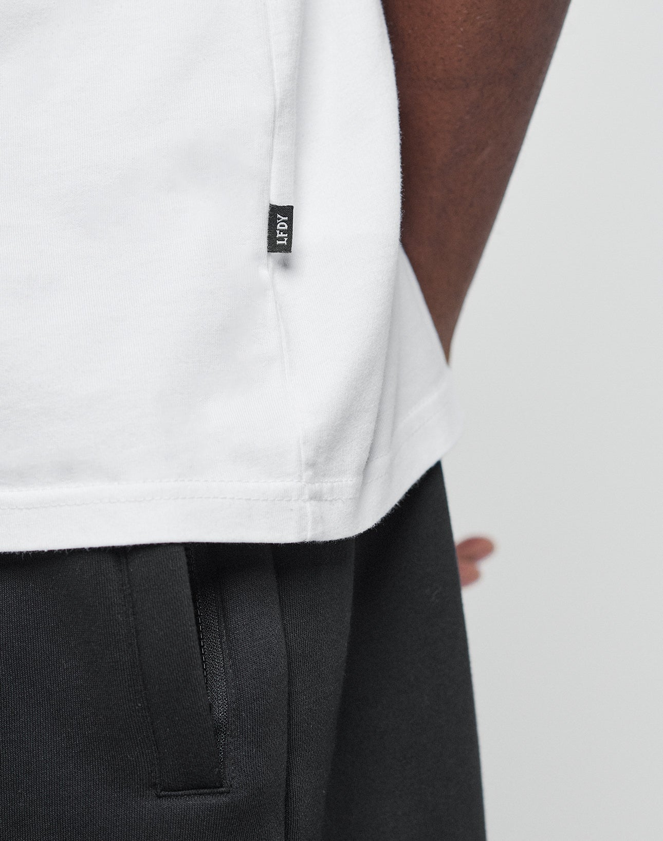 Close-up of a person wearing a white Basic LiveFast Tee by LIVE FAST DIE YOUNG, featuring a small black NOAH tag on the hem and paired with black pants. Torso, waist, and arm are shown, highlighting its Regular Fit style.