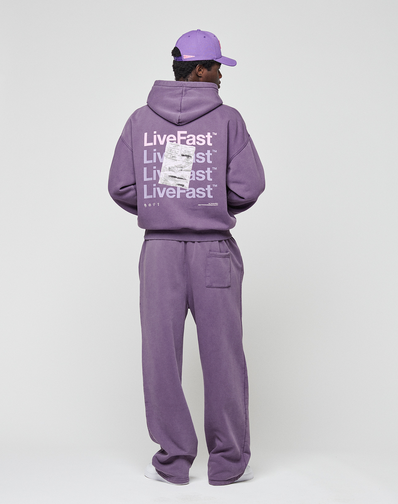 A person stands facing away in a purple LIVE FAST DIE YOUNG Basic Evidence Hooded and matching pants with a purple cap. The hoodie features repeated “LiveFast” white backprint, barcode, and small text. Background is plain light gray.