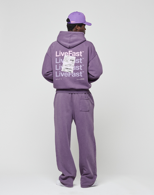 A person stands facing away in a purple LIVE FAST DIE YOUNG Basic Evidence Hooded and matching pants with a purple cap. The hoodie features repeated “LiveFast” white backprint, barcode, and small text. Background is plain light gray.