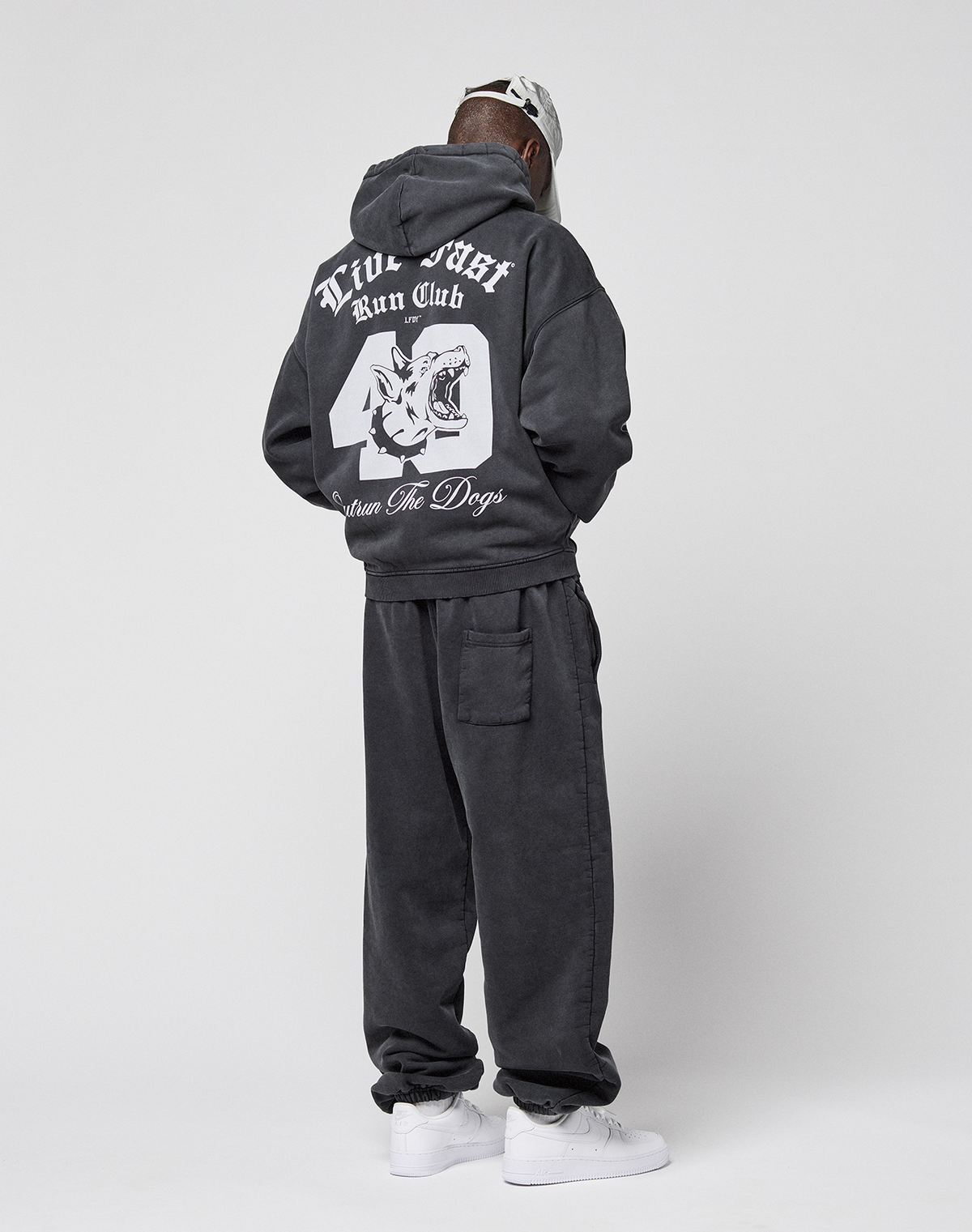 A person faces away, wearing the LIVE FAST DIE YOUNG LFDY Run Club Hooded Zip and sweatpants with a large dog head graphic and "True Issue Run Club, Down For Dogs" on the back. They pair it with white sneakers, a light cap, and an Ärmelprint.