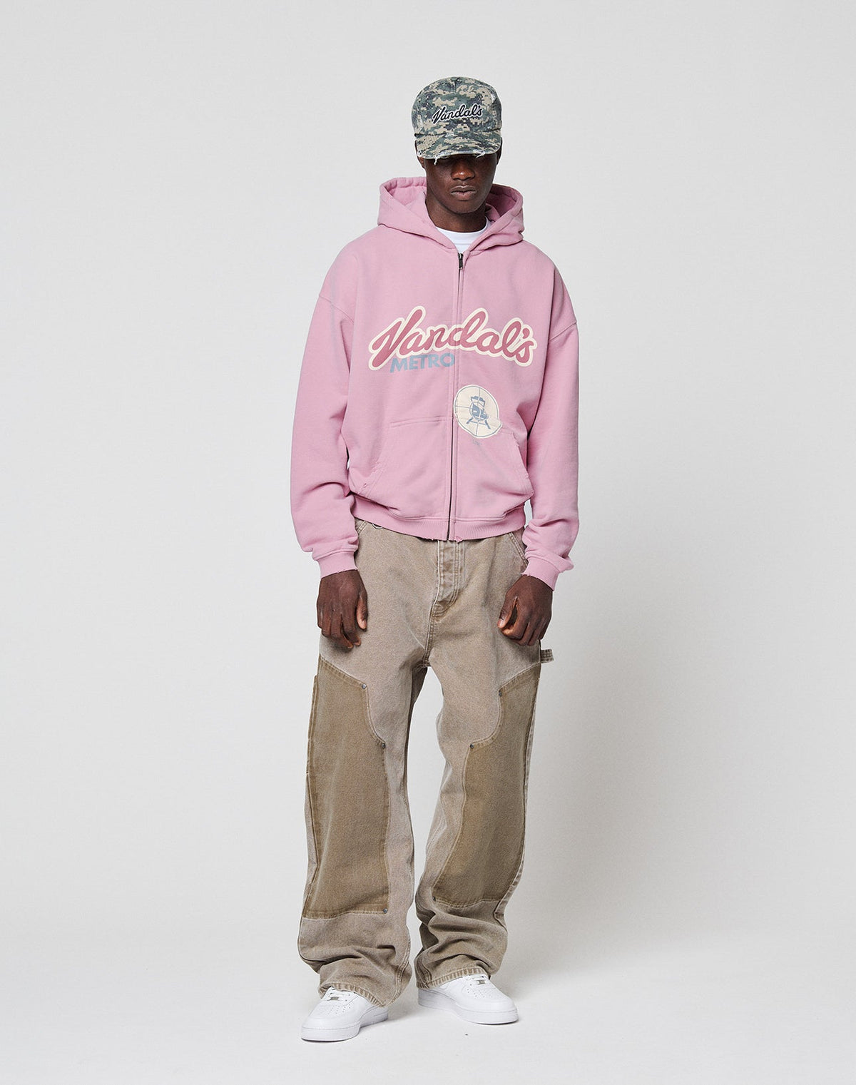 A person in a pink hooded sweatshirt with Vandals text, a camo cap, white sneakers, and LIVE FAST DIE YOUNG Faded Carpenter Pants stands against a plain white background.