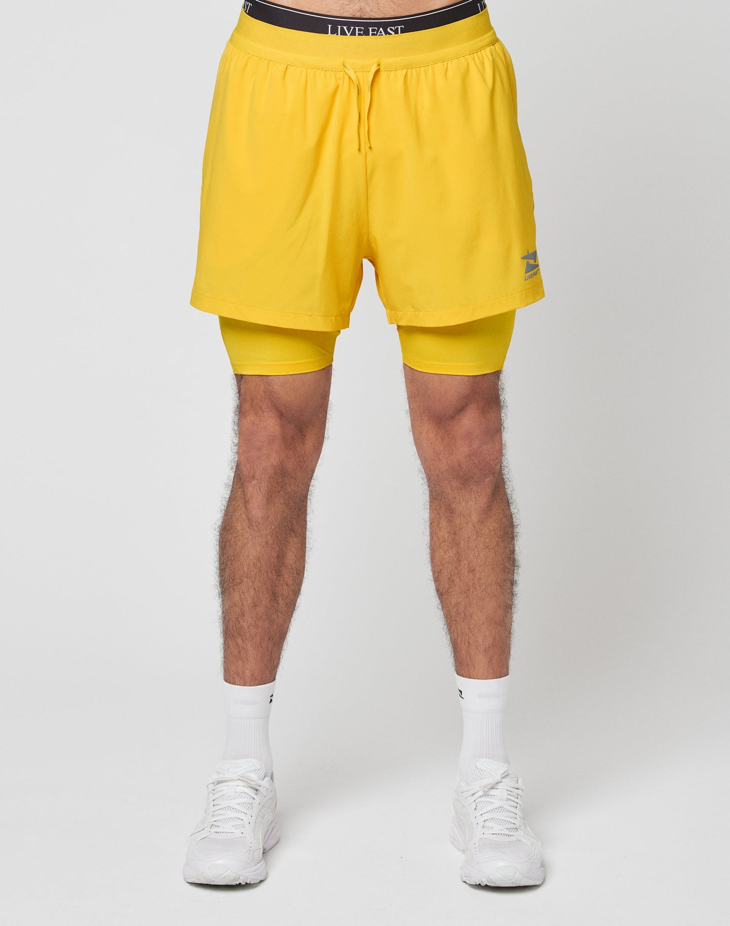 A person wearing LIVE FAST DIE YOUNG Live Fast Performance Running Shorts with integrierte Tights, white socks, and white sneakers, standing against a plain white background.