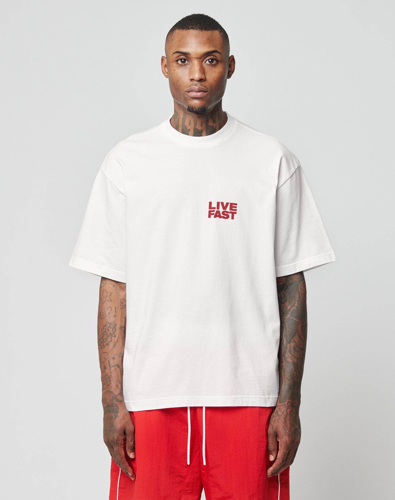 A tattooed person wears the oversize white LFDY 4 Ever Tee by LIVE FAST DIE YOUNG, featuring LIVE FAST red front and back print, paired with bright red shorts against a plain light background.