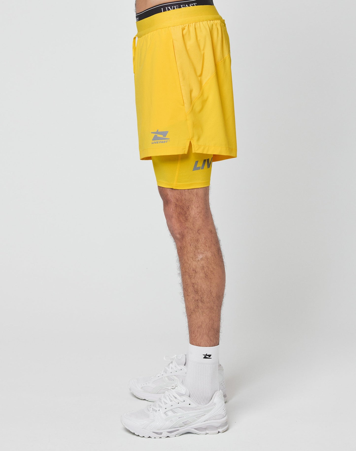 Side view of a person wearing LIVE FAST DIE YOUNG Live Fast Performance Running Shorts in yellow, featuring integrated tights, with white socks and sneakers. Only the lower half is visible against a plain white background.