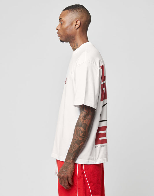 A man with short hair and arm tattoos stands in profile, wearing a LIVE FAST DIE YOUNG LFDY 4 Ever Tee and red shorts with white piping, against a plain light gray background.